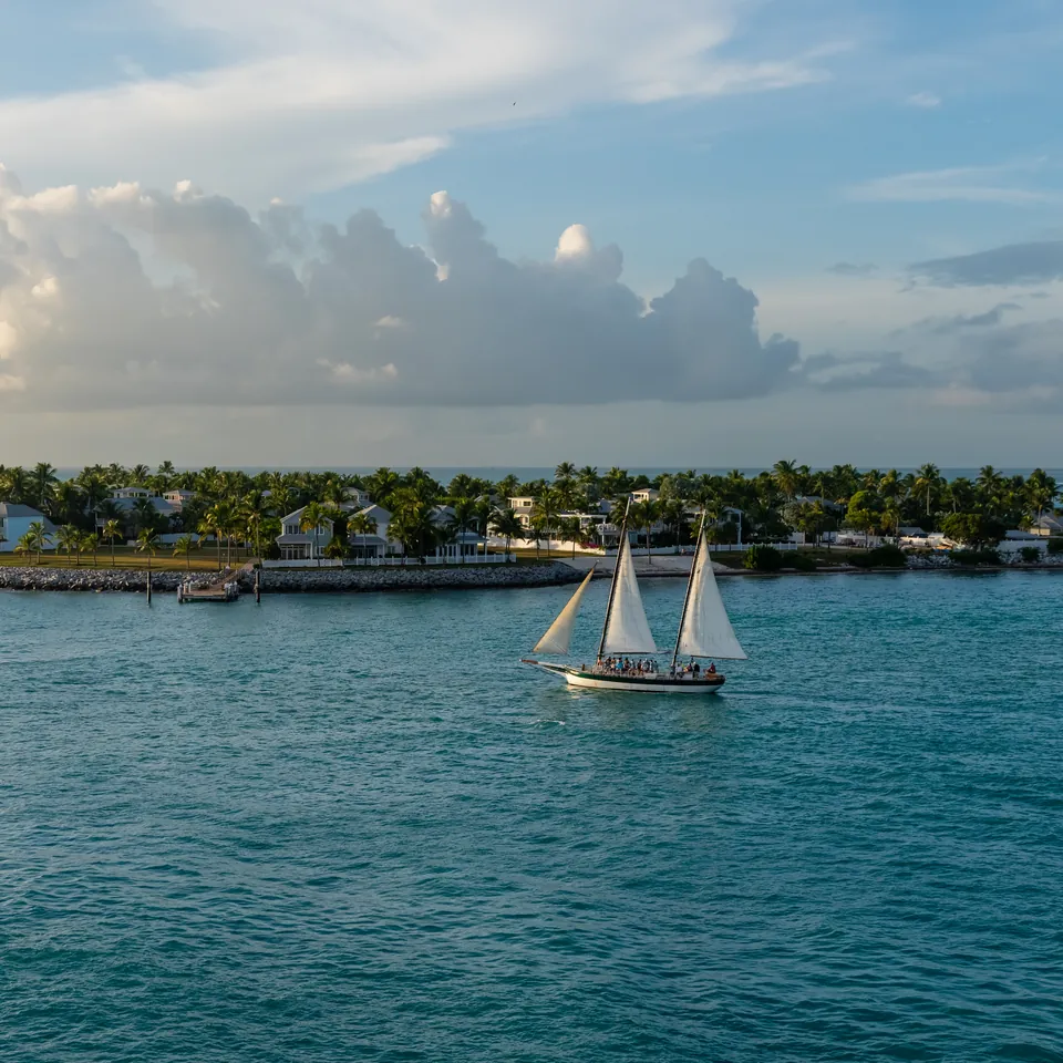 A small palm-covered island on a turquoise sea, with two white-sailed boats in front of it under scattered clouds