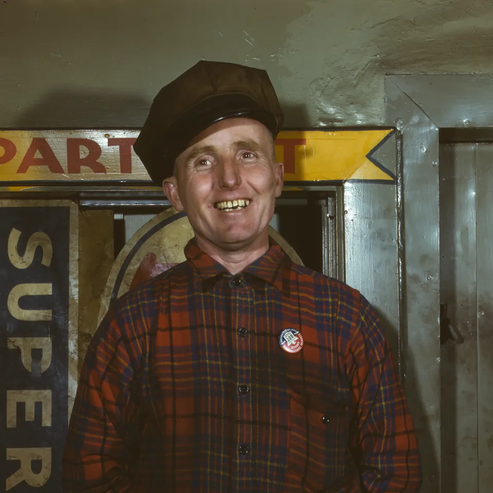 Kodachrome portrait of a grinning young mechanic in a plaid shirt and cap, standing in front of a garage sign