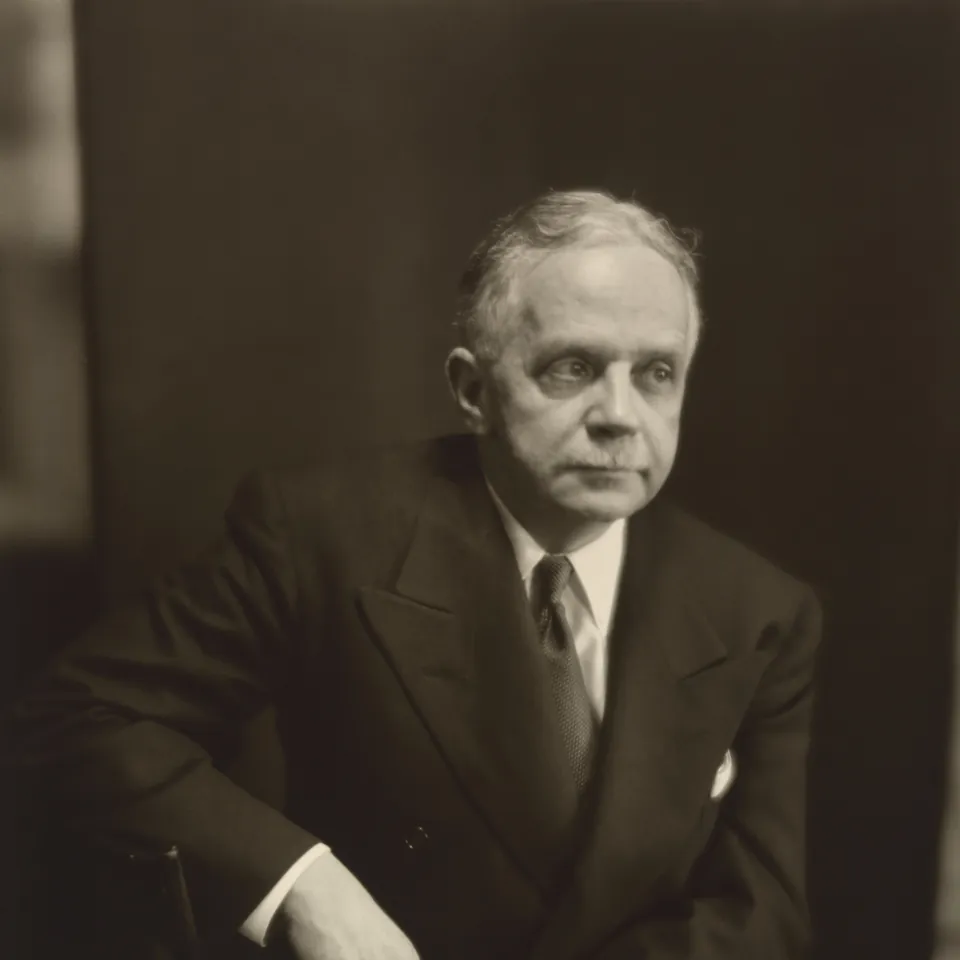 Black-and-white portrait of Walter White in a dark suit and tie, seated with his hands folded