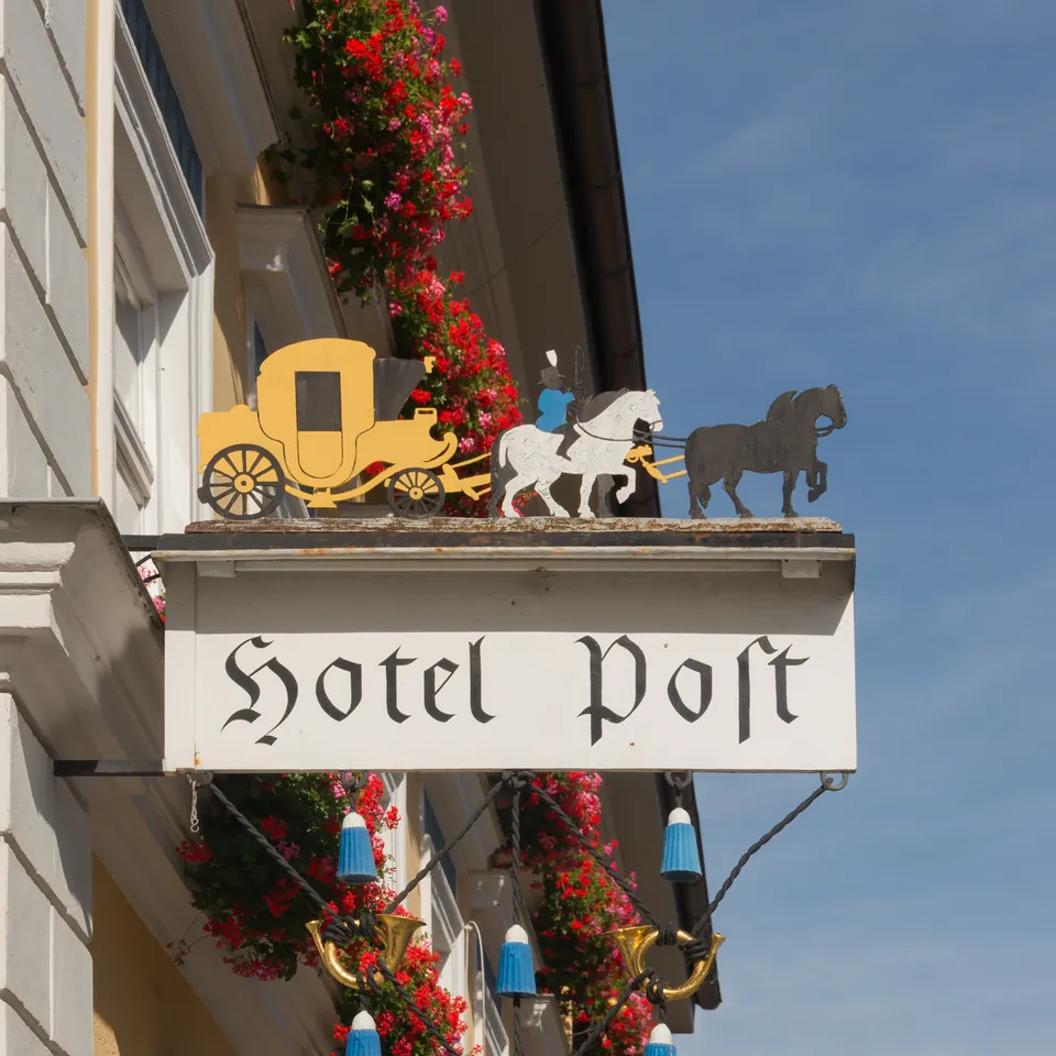 An ornate hanging inn sign reading Hotel Post in blackletter, topped by a gilded carriage and horses, beside red geraniums